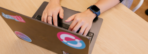 A person is typing on a laptop with colorful dozanü stickers, wearing a blue smartwatch, seated at a light wooden desk.