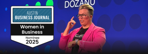 A person in a pink blazer signing on stage in front of a blue background with the word “DOZANÜ” behind them. On the left side of the image, a badge reads “Austin Business Journal – Women in Business Nominee 2025.